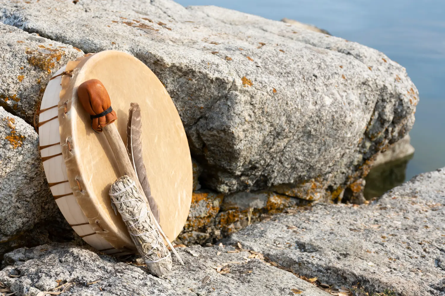 Ein Bild einer handgefertigten Meditationstrommel aus Leder, die mit einer heiligen Feder und einem Salbei-Räucherstäbchen an einer Felswand lehnt.
